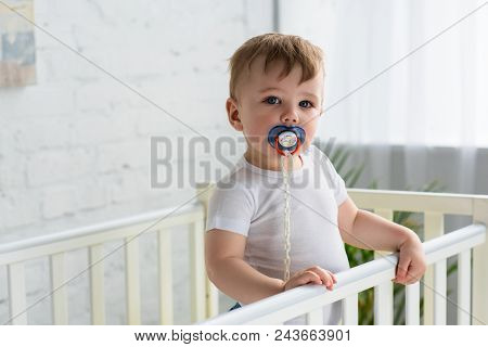 Cute Little Baby Boy With Pacifier In Baby Crib Looking At Camera At Home