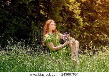 Young, Beautiful Woman In A Green Dress With Flowers In Hand Sits On A Meadow In A Mountain Forest