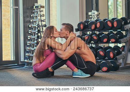 Young Attractive Sporty Couple Sitting Opposite Each Other On The Floor And Kissing At The Fitness C