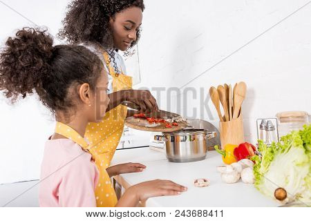 African American Mother And Daughter Putting Ingredientes In Saucepan On Kitchen