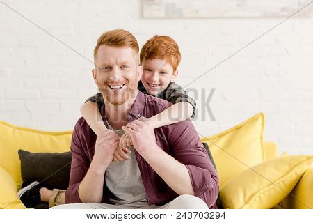 Happy Redhead Father And Son Hugging And Smiling At Camera At Home