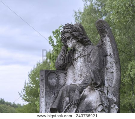 Old Stone Baroque Statue Of Sad Greving Angel Holding Head In Hand, Green Tree And Blue Sky Backgrou