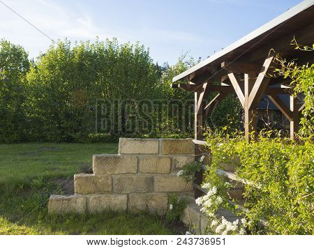 Brown Tiber Wooden Gazebo Or Pergola With Climbing Plants And White Flowers, Surrounding Sandstone W