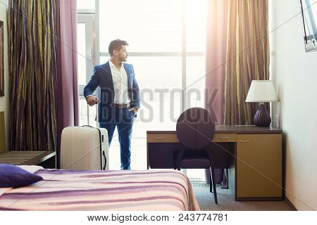 Young Businessman With Luggage In Hotel Room. Successful Man In Suit Looking At Window, Arriving To 