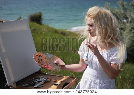 Beautiful Girl Draws A Picture In The Park Using A Palette With Paints And A Spatula. Easel And Canv