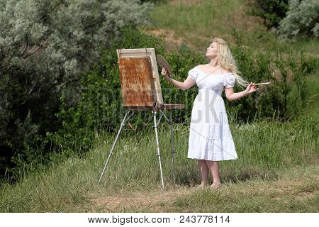 Beautiful Girl Draws A Picture In The Park Using A Palette With Paints And A Spatula. Easel And Canv