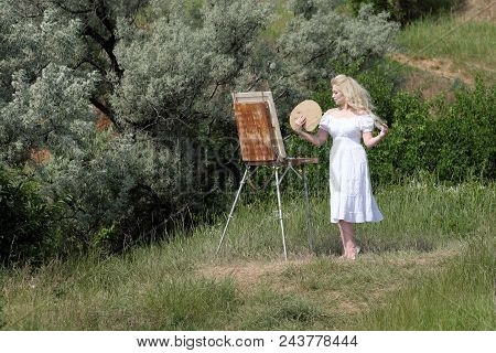 Beautiful Girl Draws A Picture In The Park Using A Palette With Paints And A Spatula. Easel And Canv