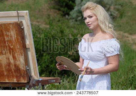 Beautiful Girl Draws A Picture In The Park Using A Palette With Paints And A Spatula. Easel And Canv