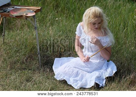 Beautiful Girl Draws A Picture In The Park Using A Palette With Paints And A Spatula. Easel And Canv