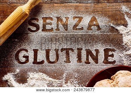 high angle view of a wooden table sprinkled with a gluten free flour where you can read the text senza glutine, gluten free in italian, next to a rolling pin and a piece of dough