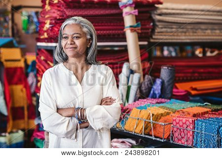 Smiling Mature Fabric Shop Owner Standing With Her Arms Crossed Surrounded By Shelves Full Of Colorf