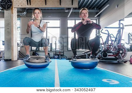 Fit Couple Working On Bosu Ball In Fitness Studio. Doing A Squat Exercise