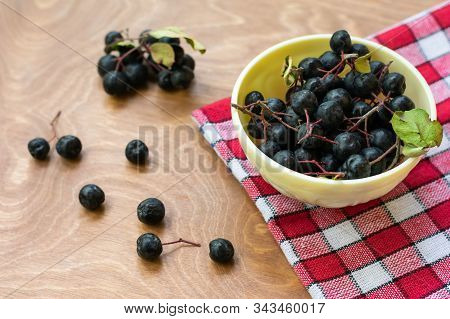 Ripe Black Chokeberries (aronia Melanocarpa) In A White Bowl On Red Napkin On Wooden Table. Bowl Ful