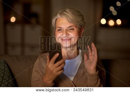 technology, old age and people concept - happy senior woman with smartphone having video call and waving hand at home in evening