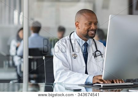 Doctor at work in office using computer with copy space in modern clinic. African happy specialist wearing white coat and sitting at desk while medical team having meeting in background.