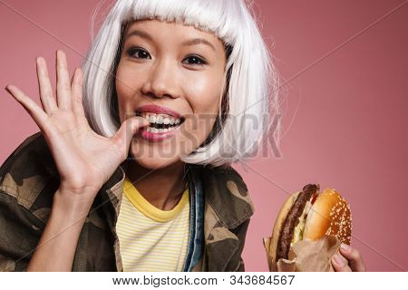 Image of young asian girl wearing white wig having fun and eating big burger isolated over pink background