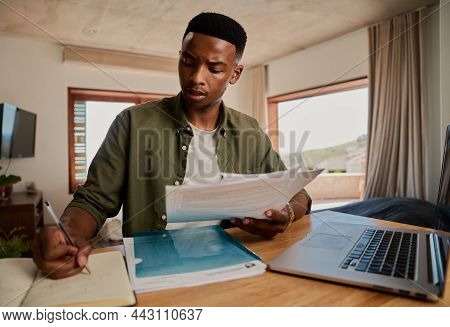 Stressed Young Adult Black Male Studying With Paper Notes, Book And Laptop In Modern Apartment.