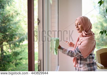Side Portrait Of A Delighted Beautiful Arab Muslim Woman With Covered Head In Hijab Washing Windows,
