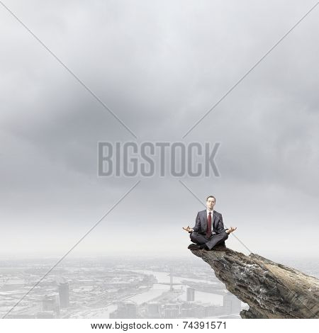 Young businessman sitting lotus pose on edge of rock mountain
