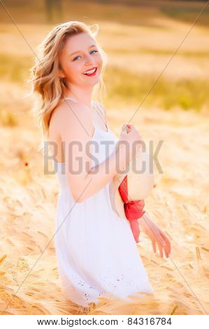 Happy Beautiful Young Blonde Girl In White Dress With Straw Hat Posing At Golden Wheat Field
