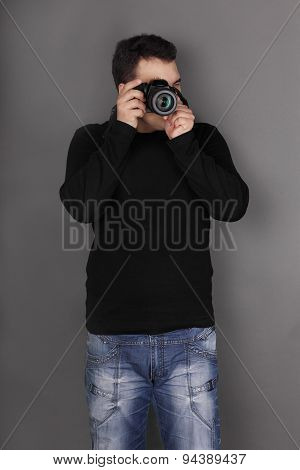 Handsome Young Man In Jeans With Modern Camera Shoots In Grey Studio