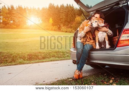 Woman with dog sits in car trunk on autumn road
