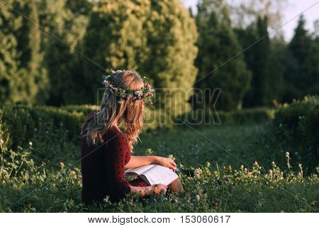 Attractive woman in wreath reading at nature, back view, copy space. Young blonde sitting with book in blooming meadow. Summer, relax, freshness, loneliness concept