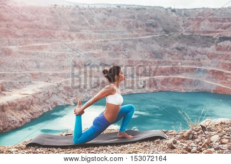 Young sporty girl wearing blue activewear stretching outdoors. Woman training yoga pose on mountain hill, calm and silent view.