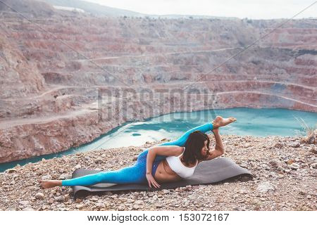 Young sporty girl wearing blue activewear stretching outdoors. Woman training yoga pose on mountain hill, calm and silent view.