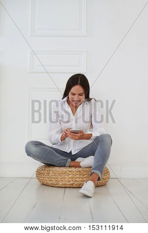 Beautiful happy Caucasian woman chatting and texting on mobile phone on the white background while sitting on a hassock