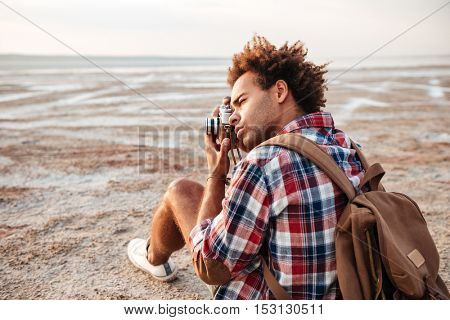 Concentrated african american young man with backpack sitting and taking pistures outdoors