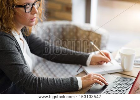 Serious business. Concentrated ginger woman working on laptop, taking notes while sitting at table near window.