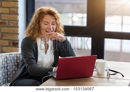 Skype conversation. Smiling red-haired woman using small modern laptop and holding her hand at her chin while sitting in cafe.
