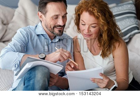Learning special aspects. Smiling middle-aged couple holding notebooks, man pointing at notes of his partner intently looking.