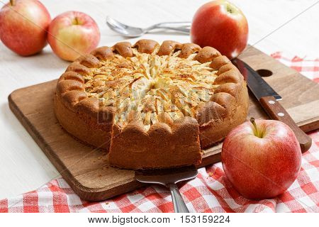 Homemade apple pie topped with slices of apples and cinnamon on white wooden table. Nearby are red apples and knife on checkered napkin.