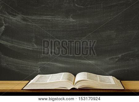 old covered text book lying opened on a blank school or college classroom desk. Blackboard background. Mockup
