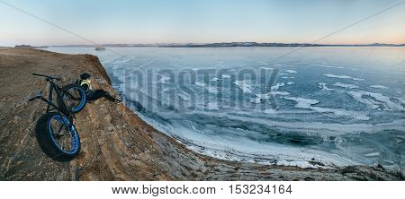 Fatbike also called fat bike or fat-tire bike - Cycling on large wheels. Traveller cyclist sits on a hillside. He is resting on a trip looking at the sunset and very beautiful frozen Lake Baikal.