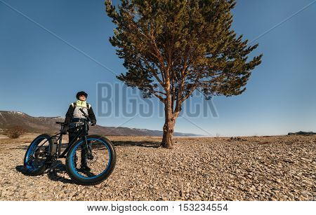 Fatbike also called fat bike or fat-tire bike - Cycling on large wheels. Traveller cyclist stands on the shore of the lake, amid picturesque frozen Lake Baikal and a huge pine tree.