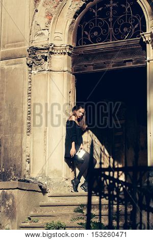 fashionable young woman or girl with pretty face in black hat and dress holding mirror and red lipstick near iron door of old ruined building on stony stairs sunny day outdoor