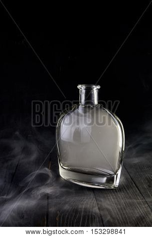 Empty glass liquid decanter graceful shape on a black wooden background with smoke in it. Crystal pitcher for alcohol in directional light. Isolated
