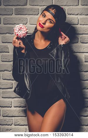 Stylish young girl in black bodysuit leather jacket and cap is holding a doughnut looking at camera and smiling on brick wall background