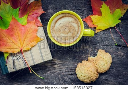 Autumn still life - Warm knitted scarf, cup of coffee, cookies and book on wooden table with fall leaves. Old book, knitted sweater with autumn leaves and coffee mug. Autumn cozy background. Top view.