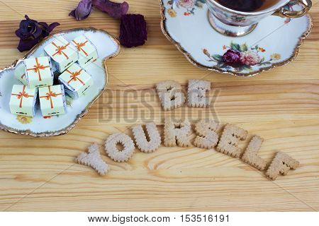 Be Yourself, inspirational slogan made from biscuit letters with chocolate sweets and a cup of tea on wooden background. Toned image.