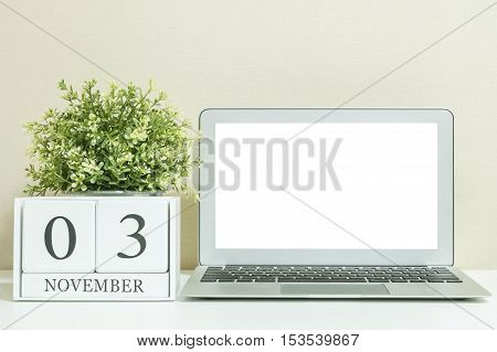 White wooden calendar with black 3 november word with white blank space at center of computer notebook on white wood desk and cream wallpaper textured background selective focus at the calendar