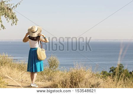 Young woman (brunette) in a blue skirt and hat looks at sea. Selective focus.