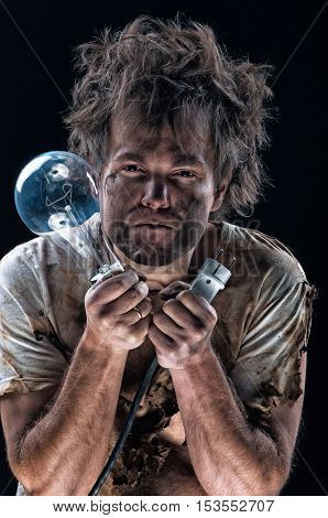 Portrait of burnt man with light bulb and electric plug over black background. Young man with dirty burnt face in funny sad expression in electricity DIY has a electric shock.