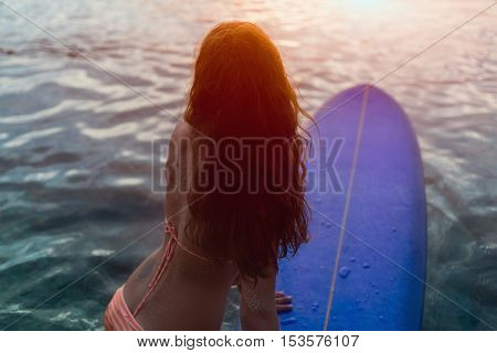 Rear view of beautiful sexy young surfer girl in bikini with blue surfboard on a beach at sunset or sunrise