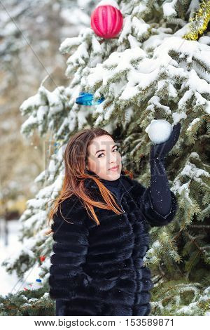 Portrait of pretty attractive girl in fur coat and gloves in the winter park. She decorates the Christmas tree balls. Winter fun. Walks in the open air. Christmas mood.