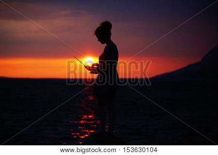 pretty sexy female silhouette of woman or girl with computer or laptop outdoor over dramatic dark sky with clouds and sea or ocean water horizon on evening or twilight on natural sunset background