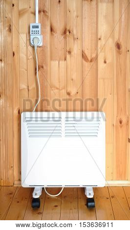 Working white electric convector heater plugged to timer power socket operated in smart house system against wooden wall inside room vertical front view closeup